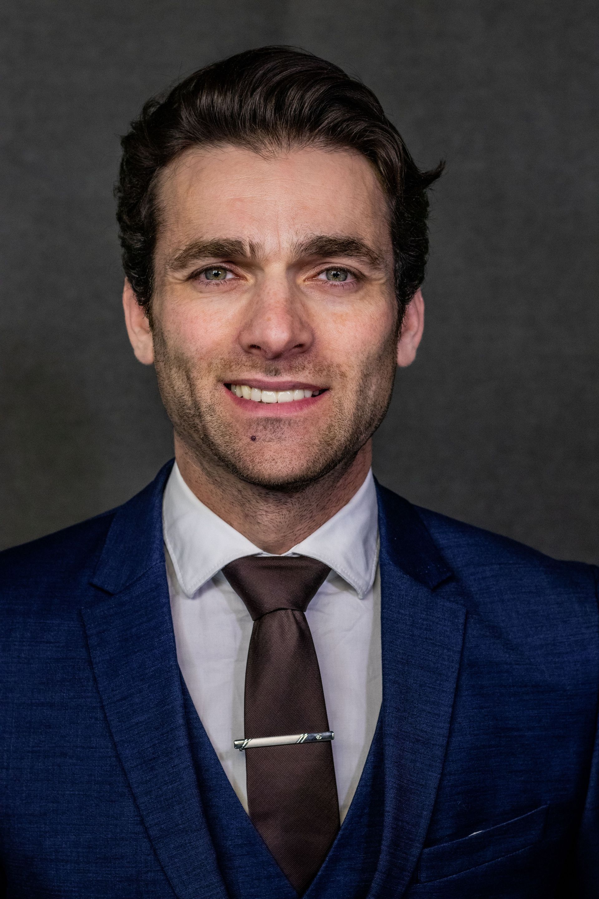 Studio portrait of a man in a blue suit, white shirt, and brown tie, smiling against a dark background