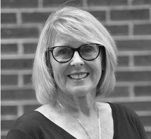 A black and white photo of a woman wearing glasses and smiling in front of a brick wall.