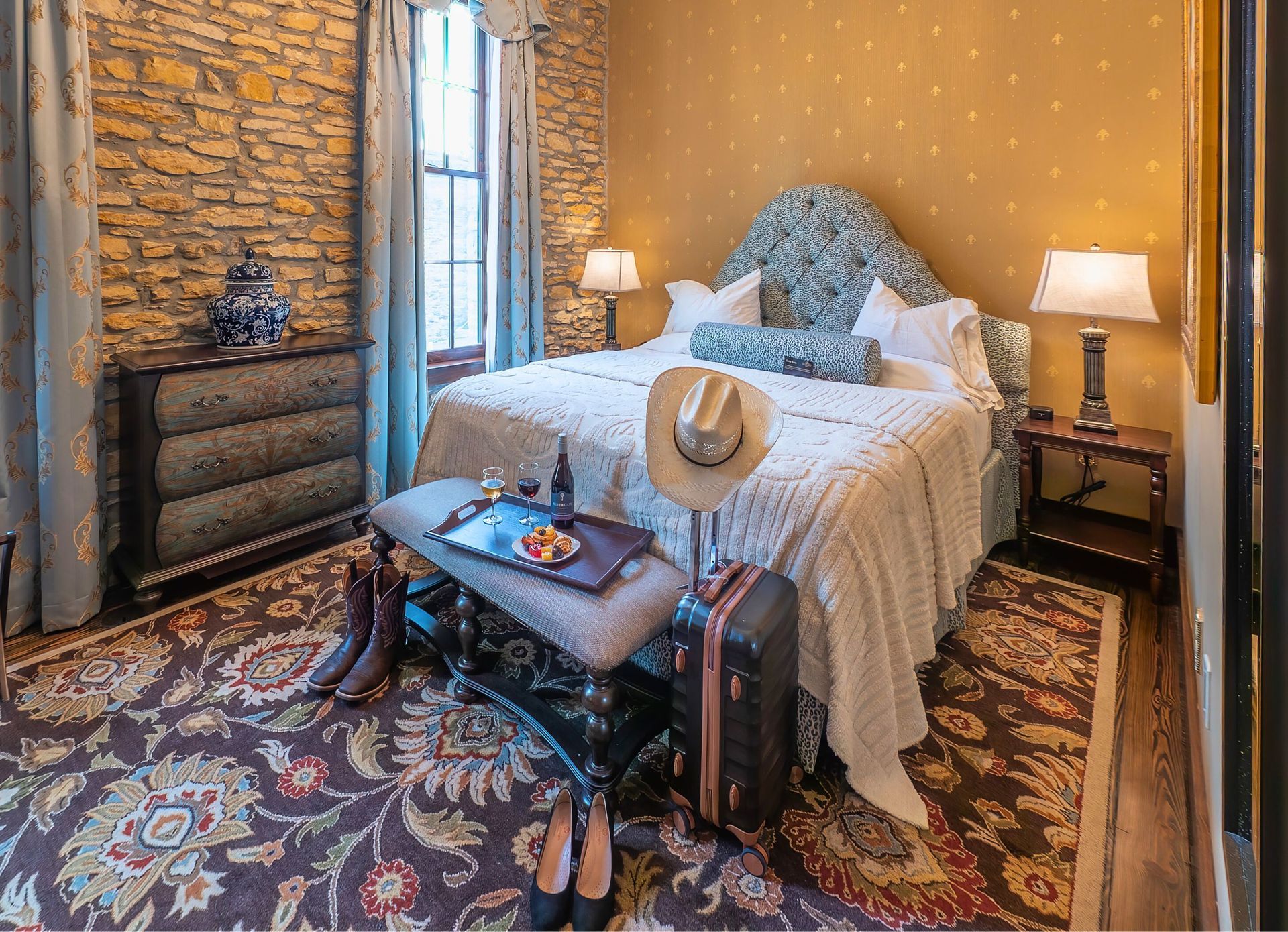 A luxury hotel bedroom with a stone wall, patterned rug, king bed, vintage suitcase, and a cowboy hat on a bench.