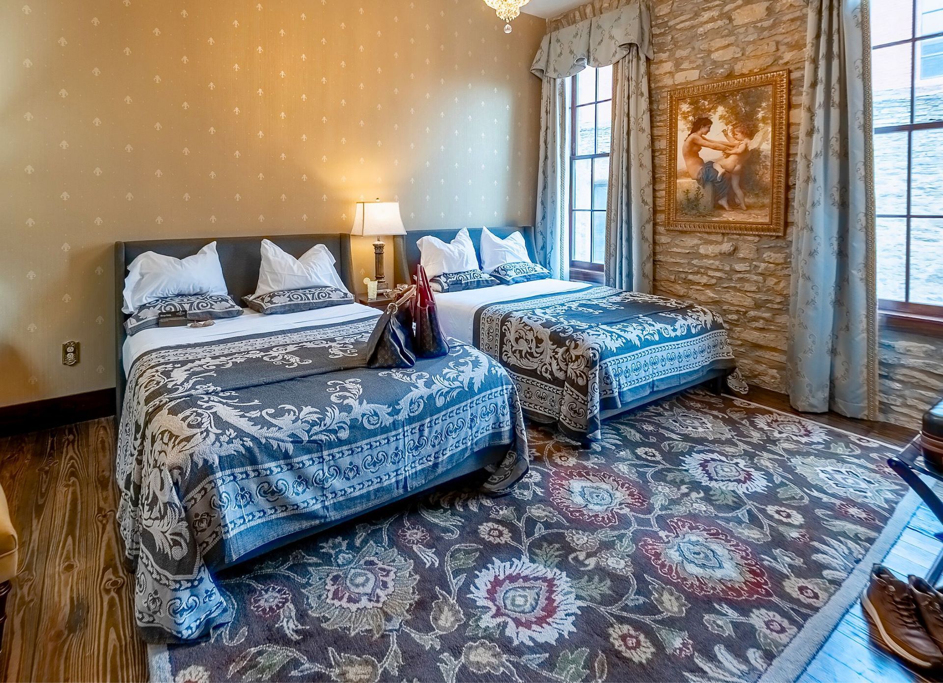 A bedroom with two beds featuring blue patterned quilts, a stone wall, floral rug, and a framed painting.
