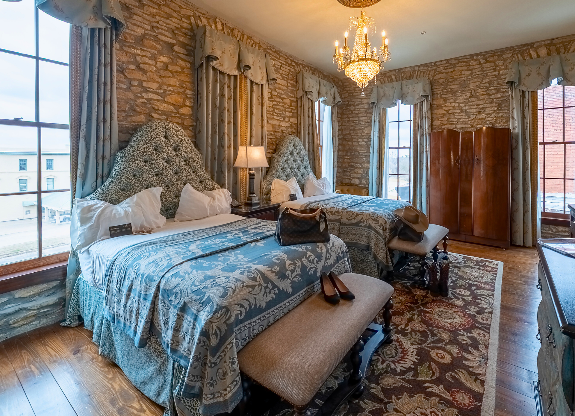 A bedroom with two beds, ornate blue and white floral bedding, patterned wallpaper, a crystal chandelier, and wood floors.
