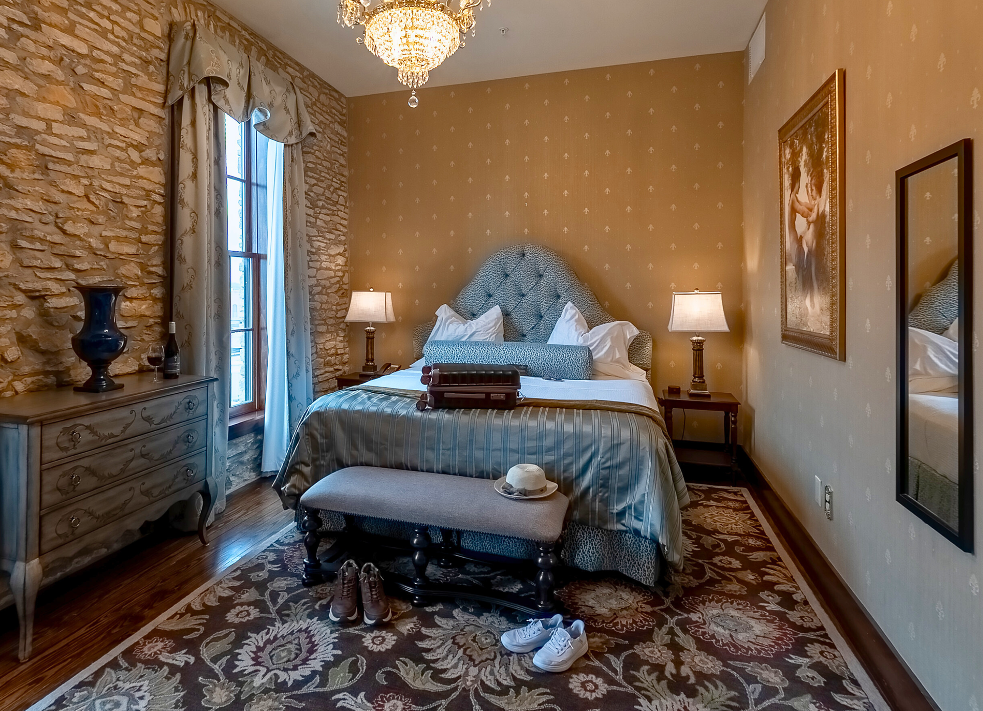 A furnished bedroom with a stone wall, floral bed frame, patterned rug, and a mirror on a gold wall.
