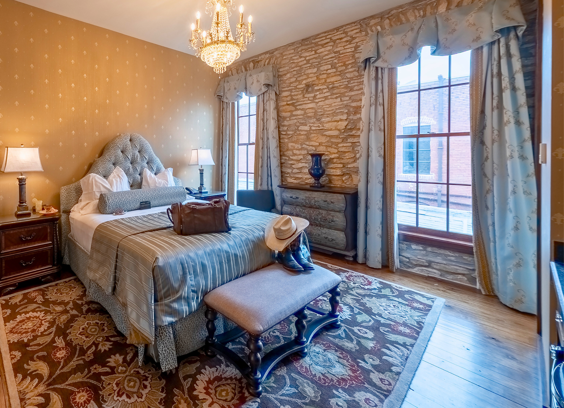 A luxury bedroom with a queen bed, stone wall, chandelier, patterned rug, and large windows with blue draped curtains.