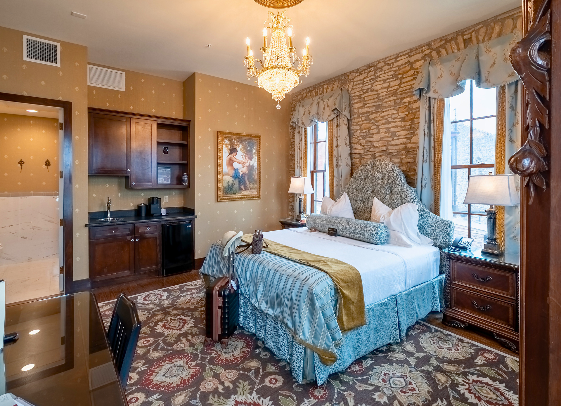 A hotel suite with a bed, chandelier, kitchenette, and patterned rug, featuring gold, blue, and dark wood tones.