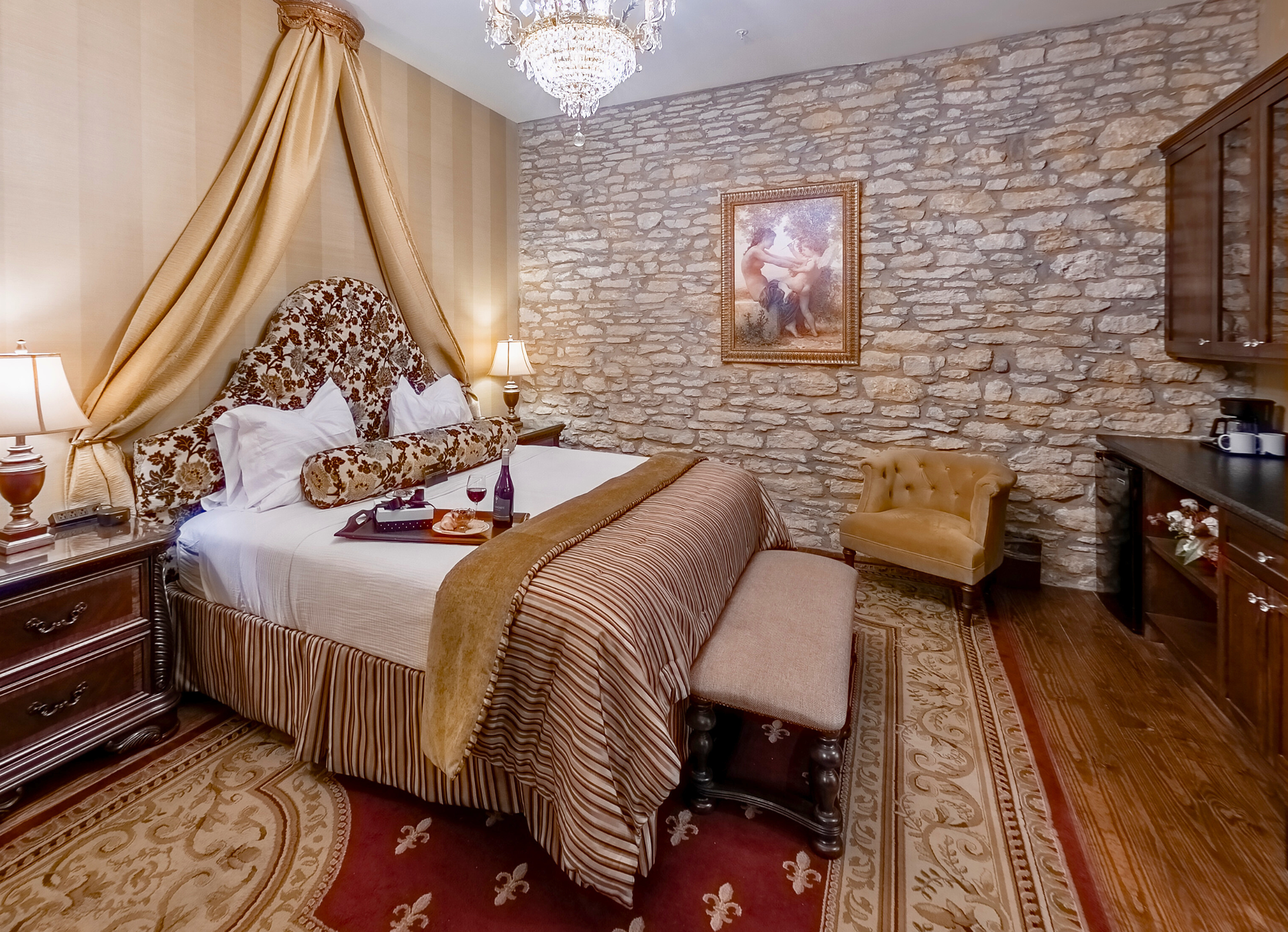 A hotel bedroom featuring a bed with a gold canopy, a stone feature wall, a velvet chair, and ornate patterned rugs.