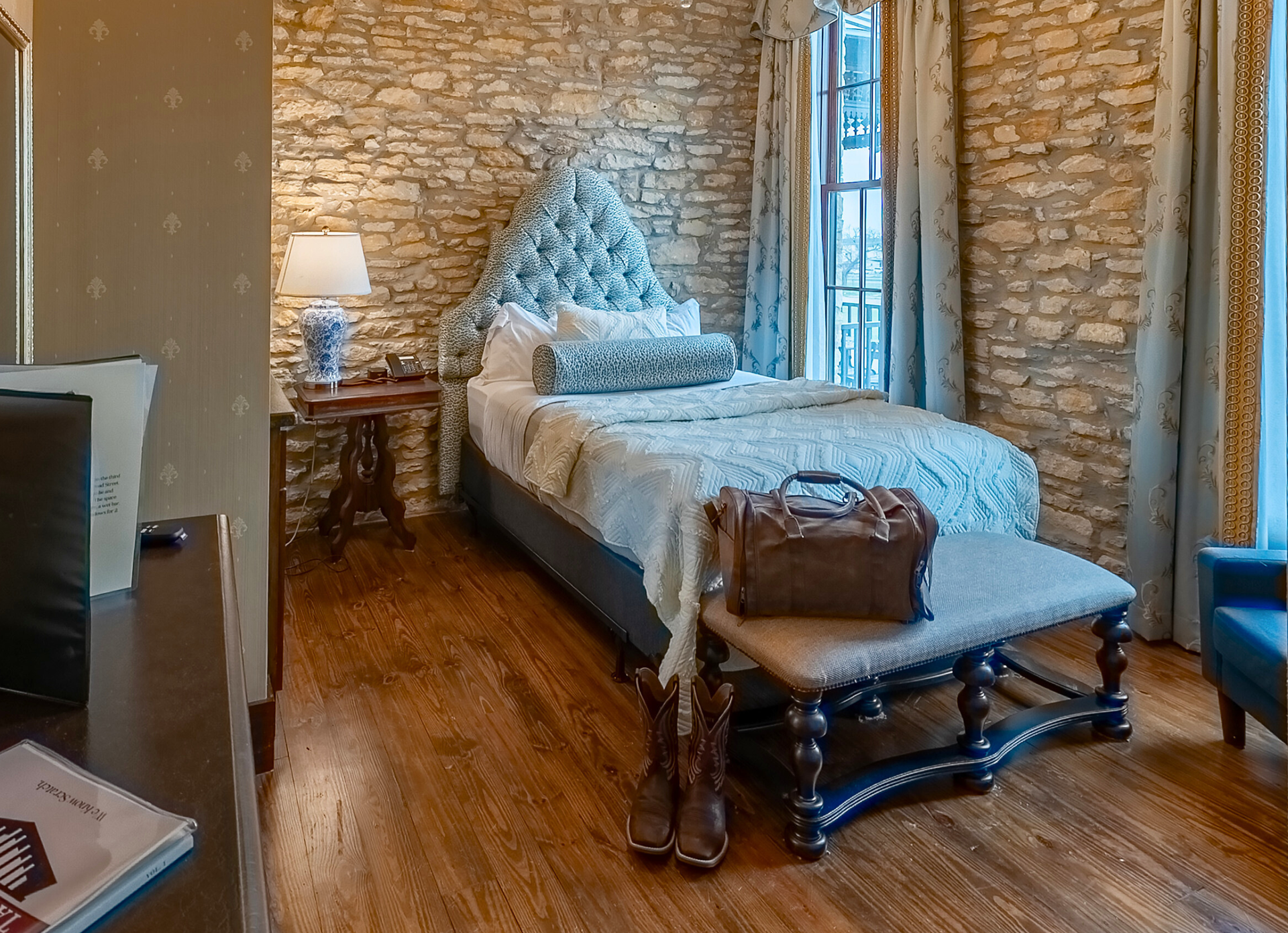 A guest room with a stone wall, a blue tufted headboard on a bed, and a leather duffel bag on an upholstered bench.