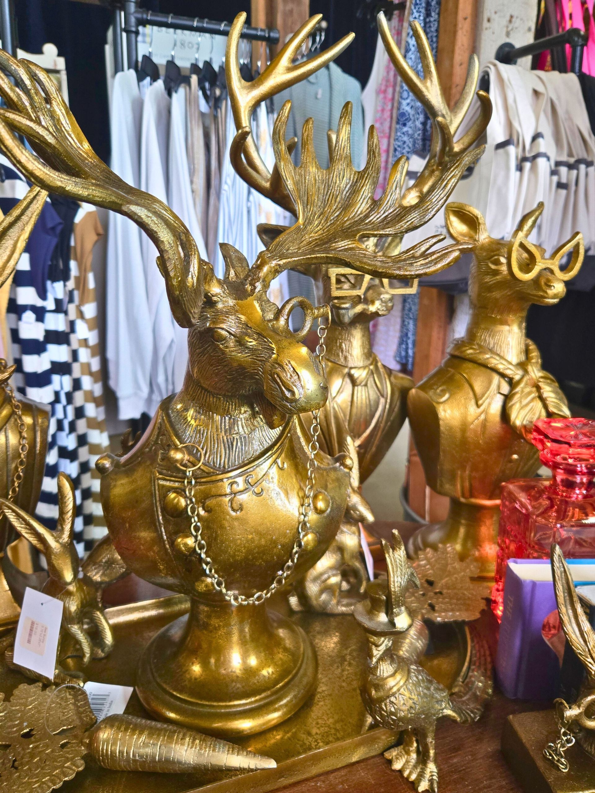 An antique store displays a wooden table holding brass figures, with racks of clothing and shelves of items nearby.