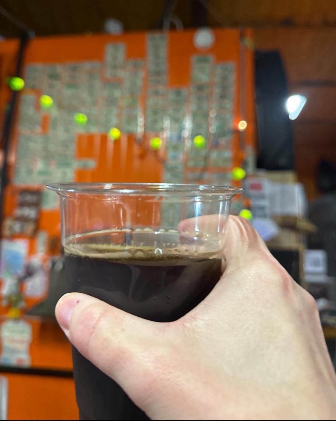 A hand holds a clear plastic cup filled with a dark liquid in front of an orange wall covered in taped paper notes.