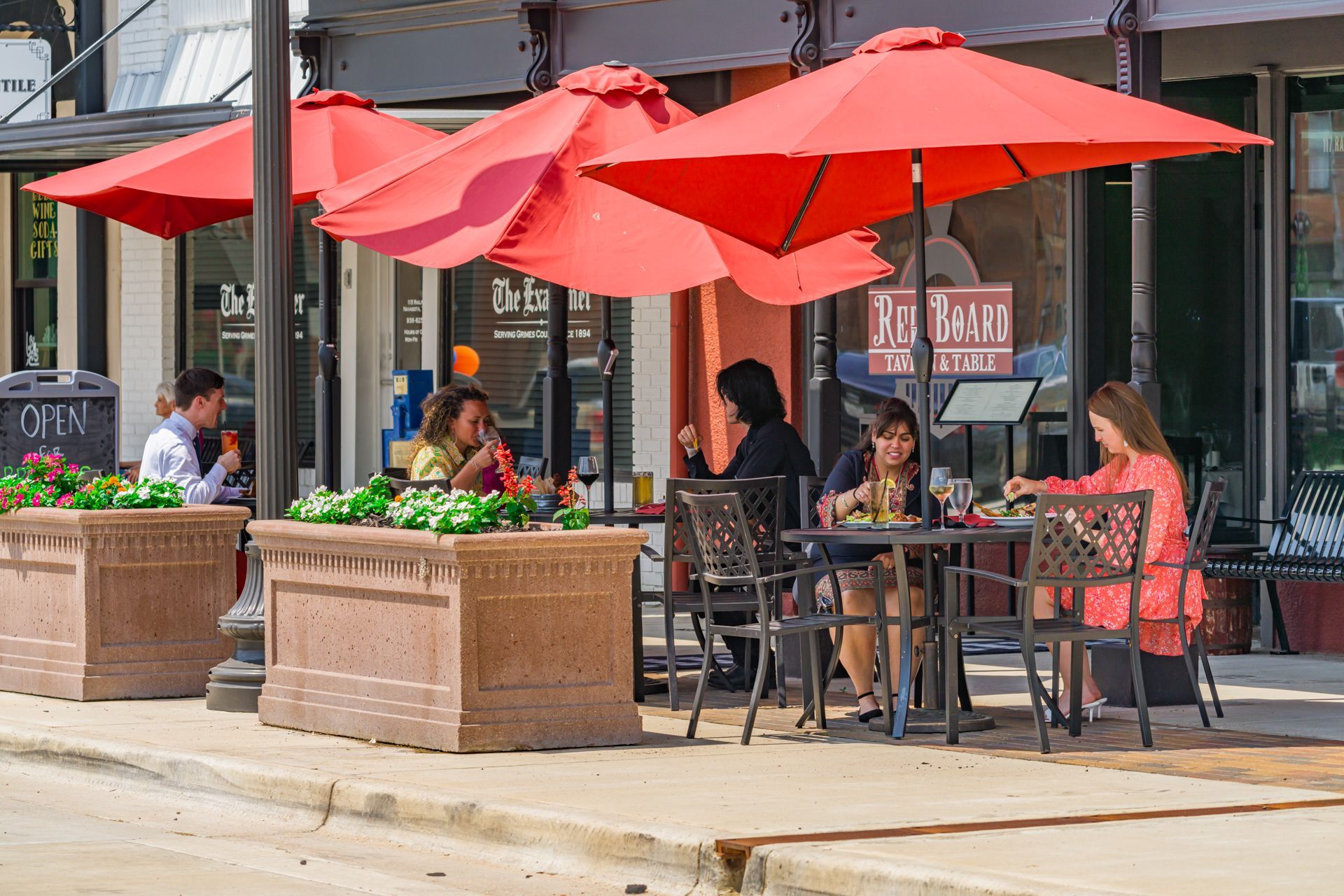 Patrons dine at outdoor tables shaded by large red umbrellas in front of a building with a storefront sign.