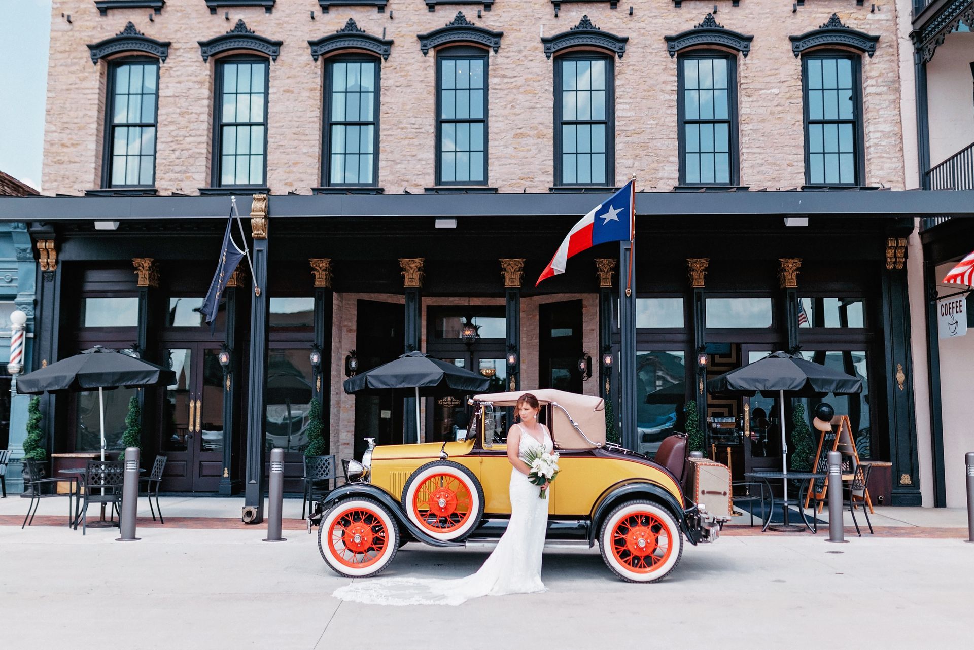 A bride in a white gown stands beside a vintage yellow car in front of a multi-story brick building with black trim.