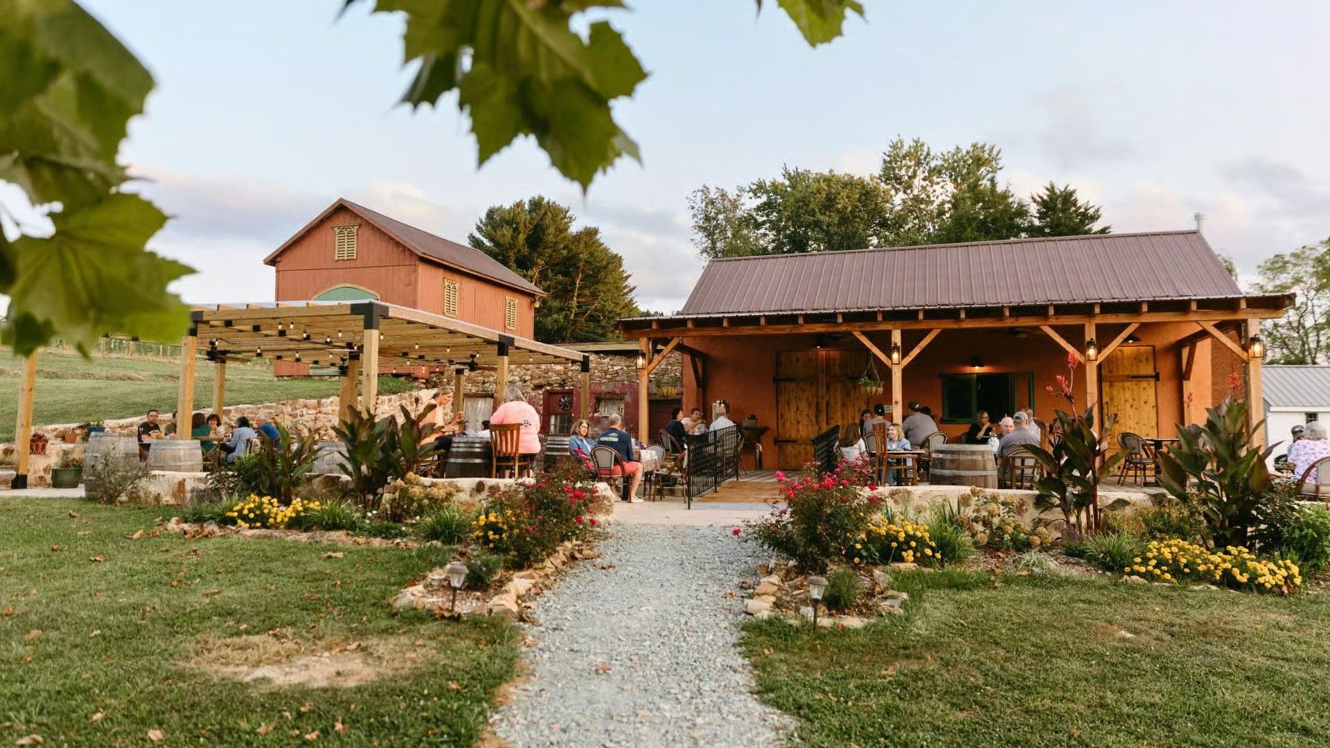 People gather on the patio and lawn of a rustic, barn-style winery building at sunset.