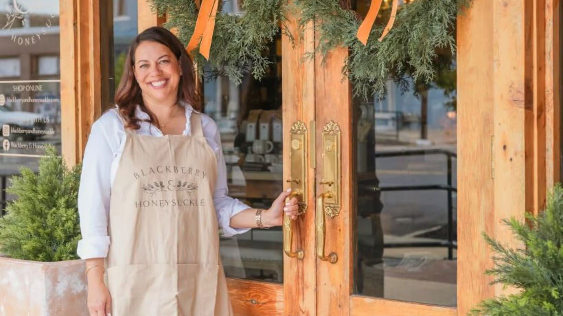 A smiling person wearing a tan apron stands in front of a wooden double door decorated with a greenery wreath.