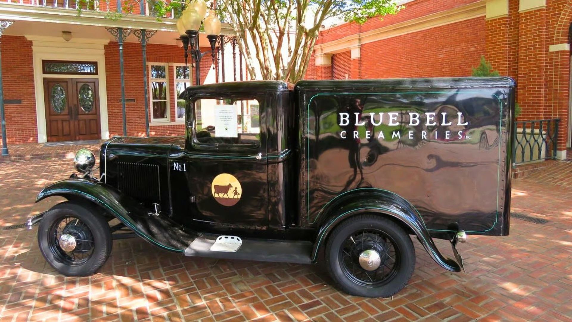 A black vintage Blue Bell Creameries delivery truck parked on a brick patio in front of a brick building.