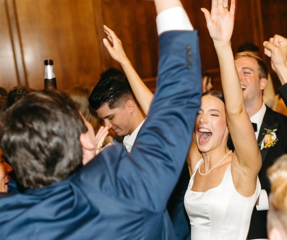 A bride and guests celebrate with hands raised and joyful expressions at an indoor wedding reception.