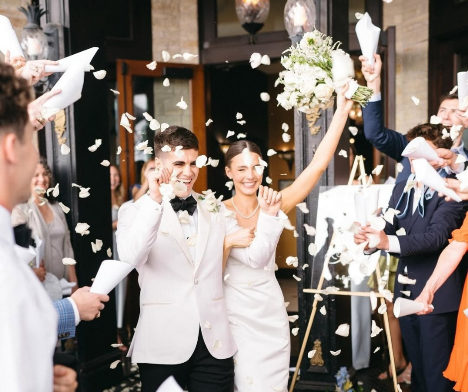 A smiling couple in wedding attire walks outside as guests throw white petals, capturing a joyous exit.