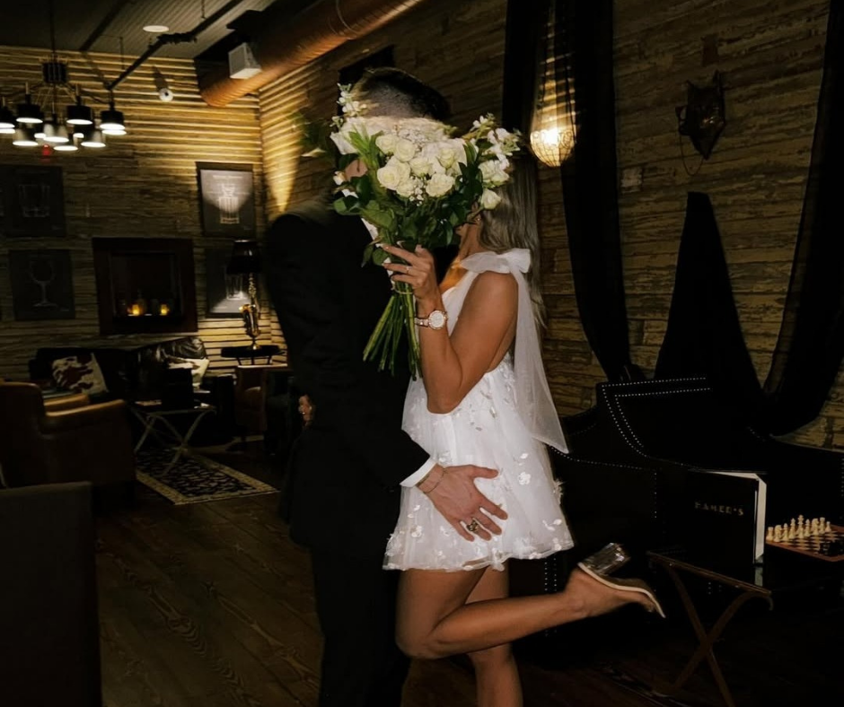 A couple embraces in a dimly lit, rustic room, hiding their faces behind a large bouquet of white roses.