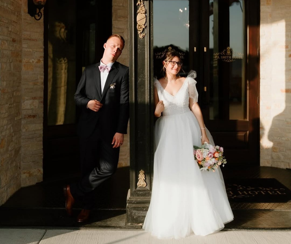 A couple in wedding attire stands on either side of a dark pillar in front of an entrance at sunset.