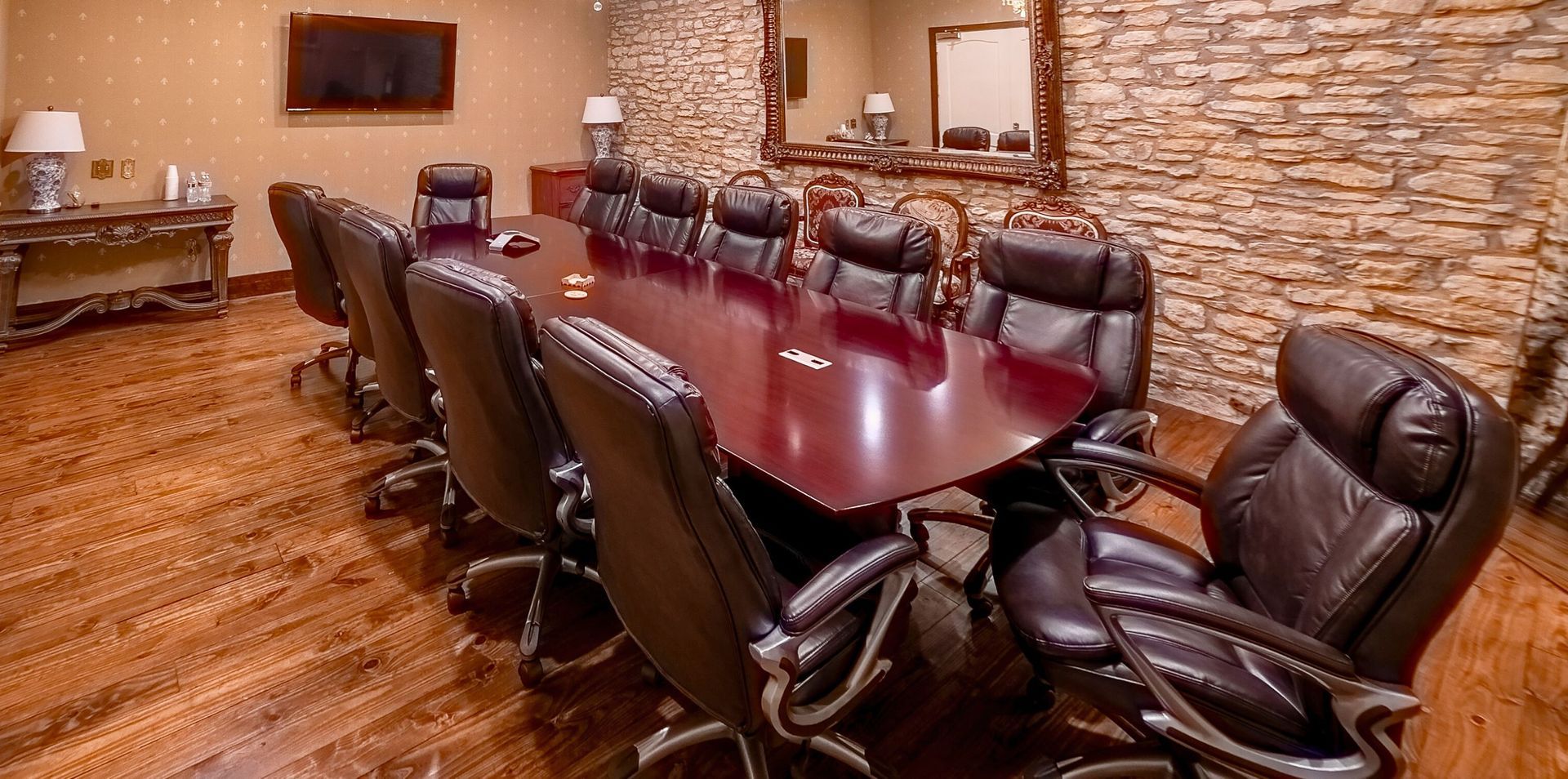A long boardroom table with leather chairs on a wood floor, featuring a stone wall, mirror, and television screen.
