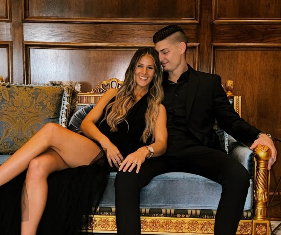 A couple in formal black attire sitting together on a blue and gold vintage-style sofa in front of wood-paneled walls.