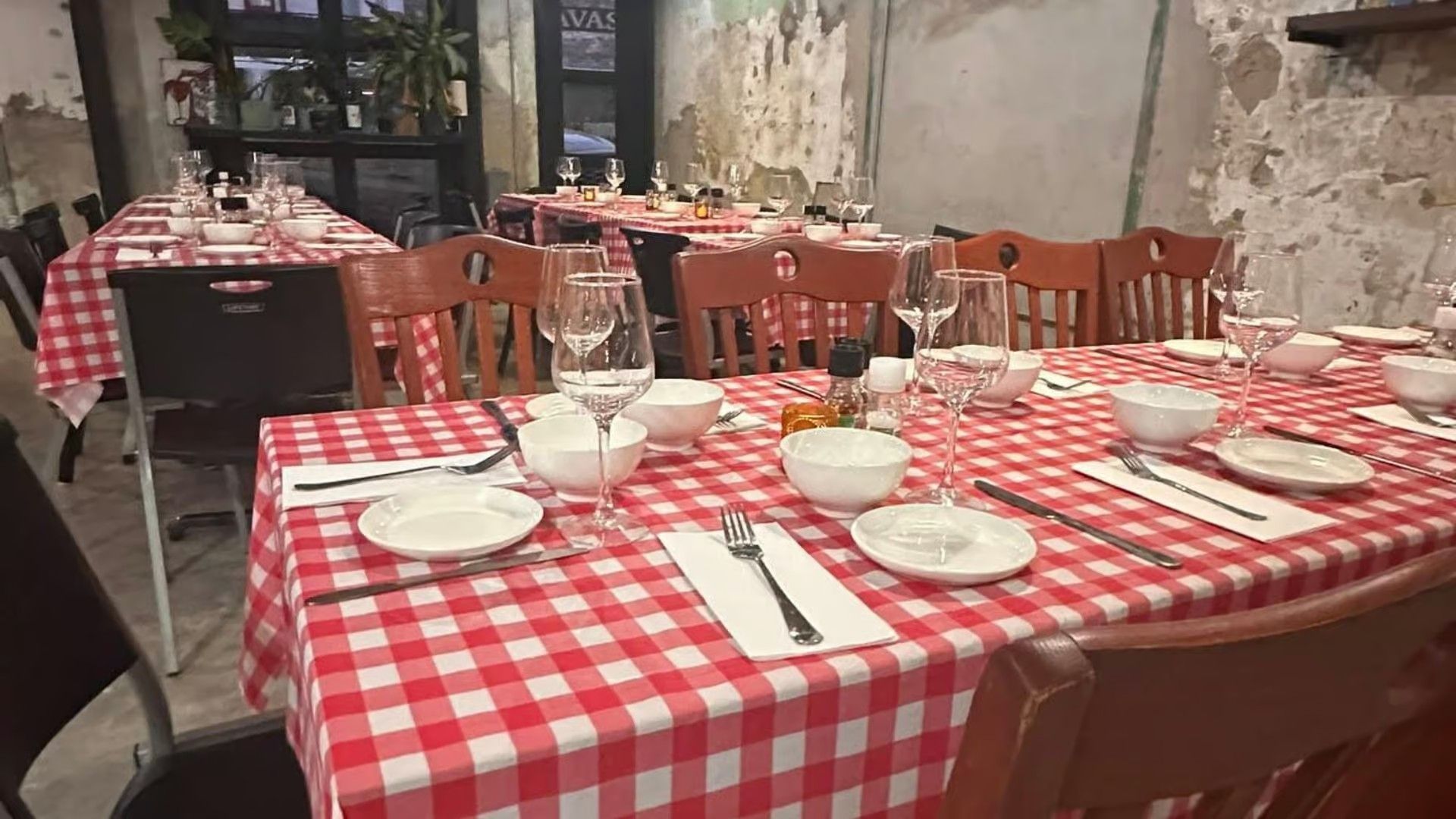 A restaurant dining room with tables covered in red and white checkered cloths, set with bowls, plates, and wine glasses.