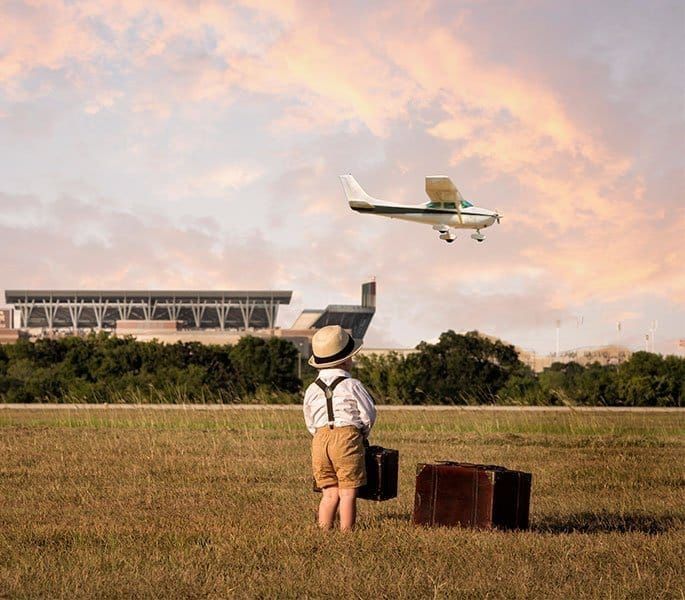 A child in vintage clothing with two suitcases stands in a field looking at a small airplane flying overhead.