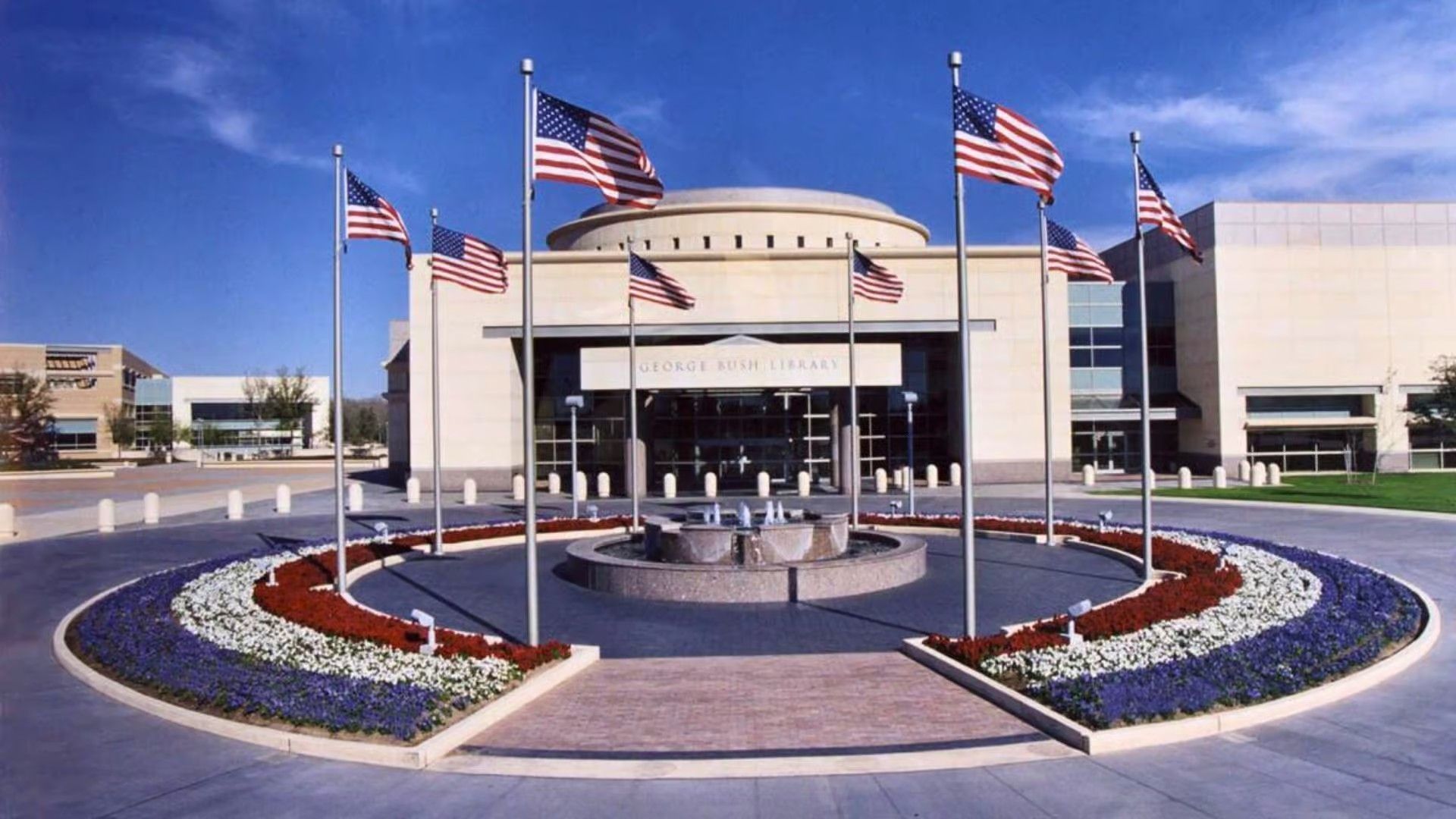 The George W. Bush Presidential Center with a circular flower bed and American flags under a bright blue sky.