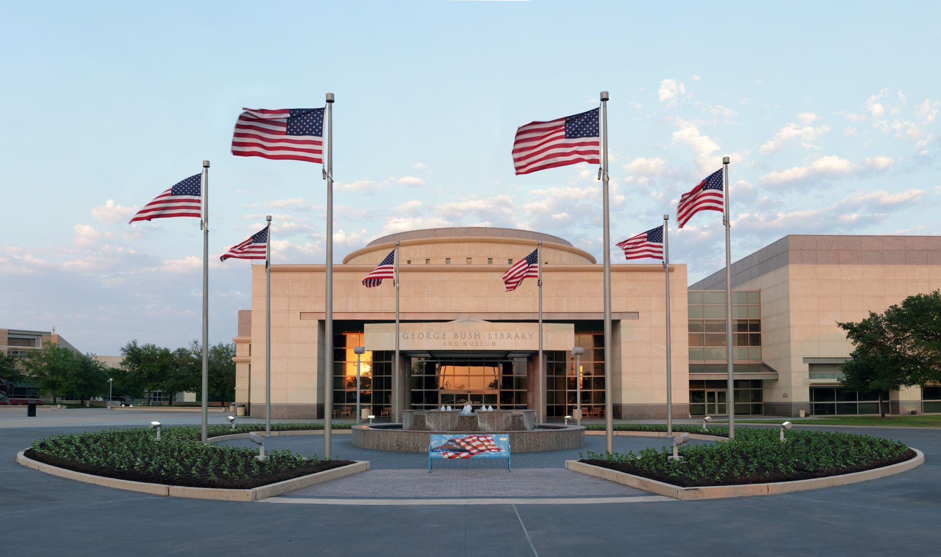 The George W. Bush Presidential Center with a circular flower bed and American flags under a bright blue sky.