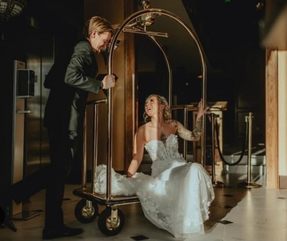 A person in a suit pushes a gold luggage cart carrying a person in a white wedding gown inside a dimly lit hotel lobby.