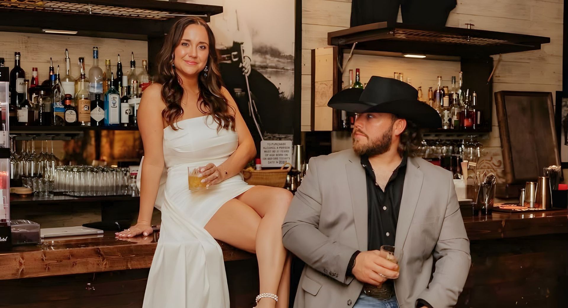 A woman in a white dress and a man in a gray suit and cowboy hat sit at a bar, each holding a glass.
