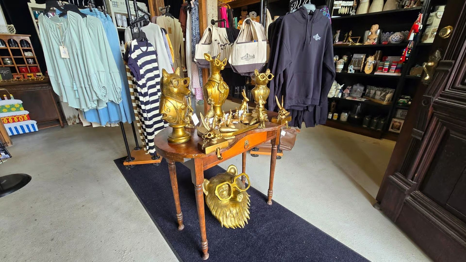 An antique store displays a wooden table holding brass figures, with racks of clothing and shelves of items nearby.
