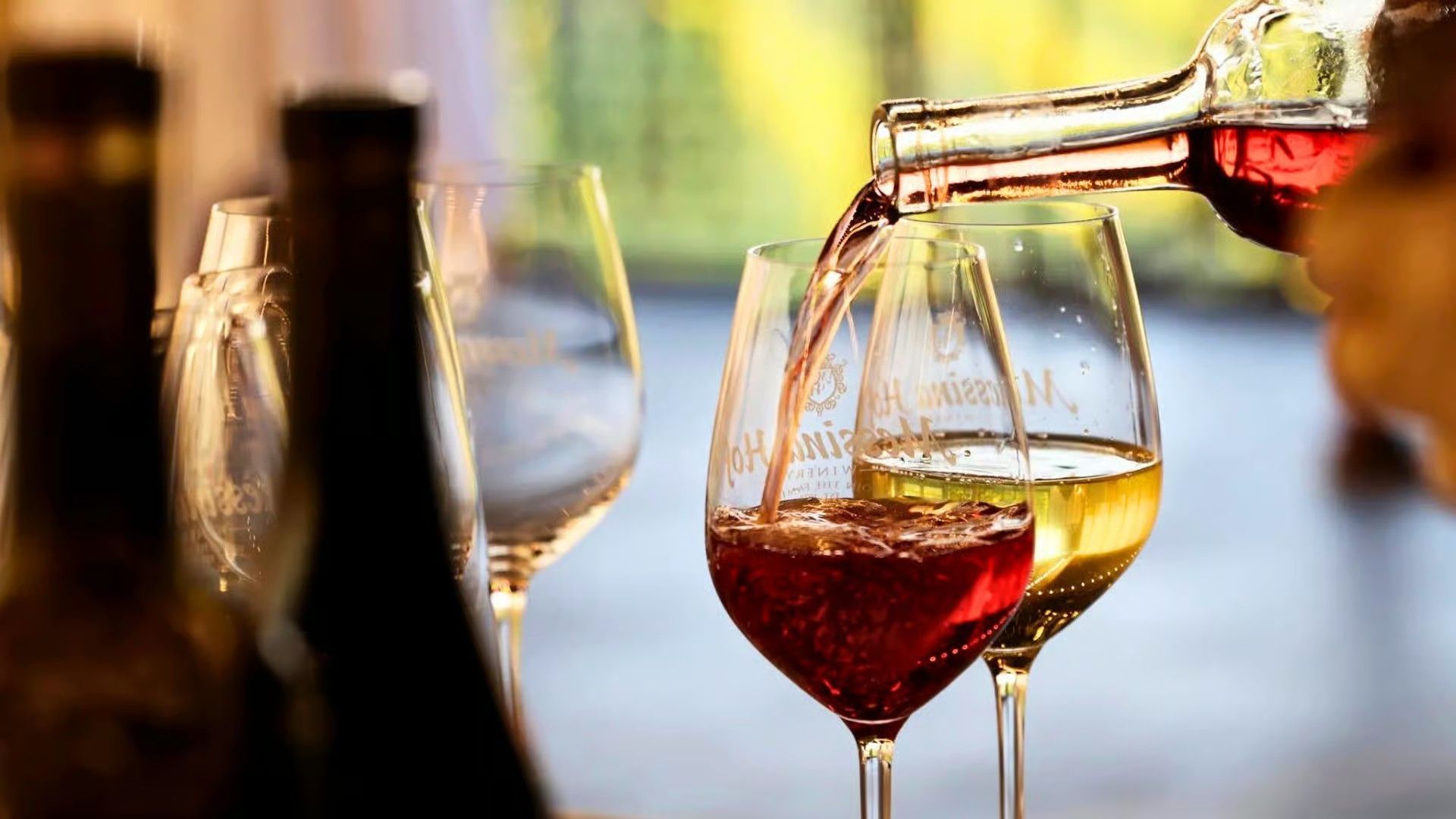 Red wine being poured into a wine glass, with other glasses and a bottle visible in the background.