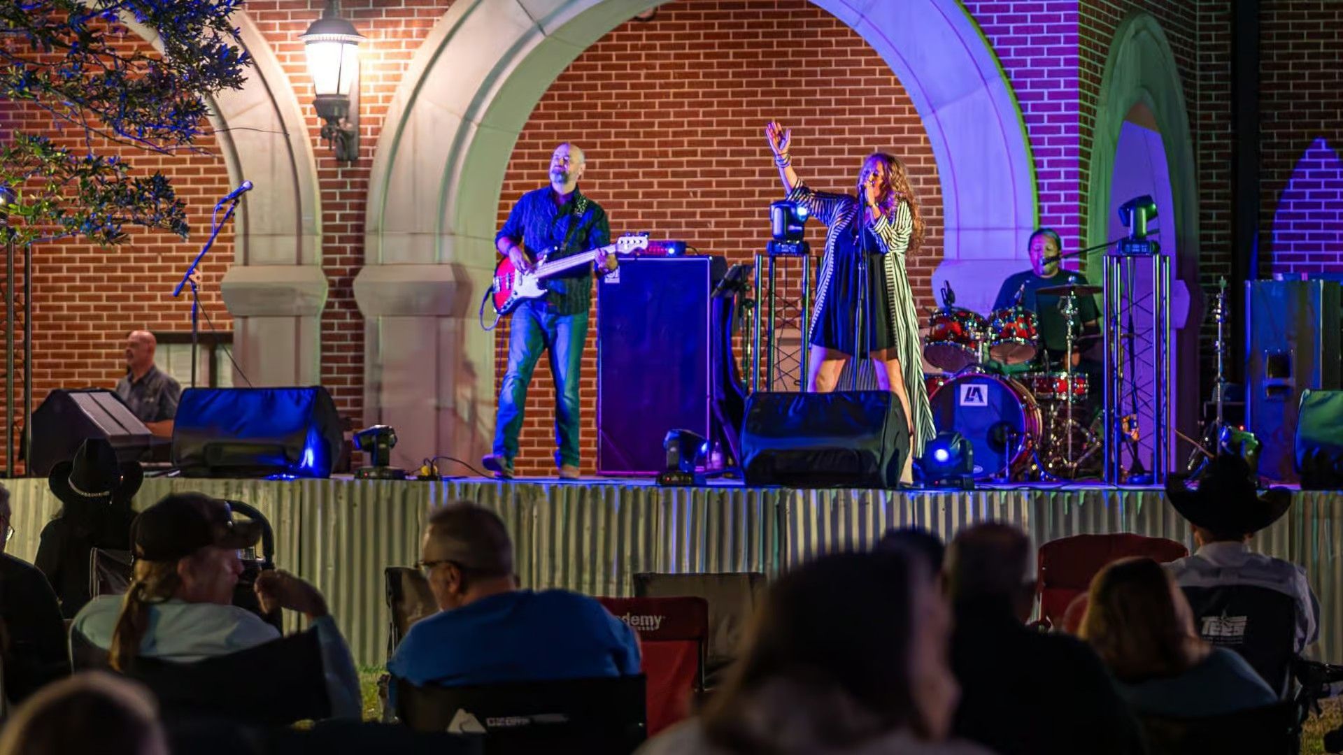 A band performs on an outdoor stage at night in front of a brick building with arched entryways and a seated audience.