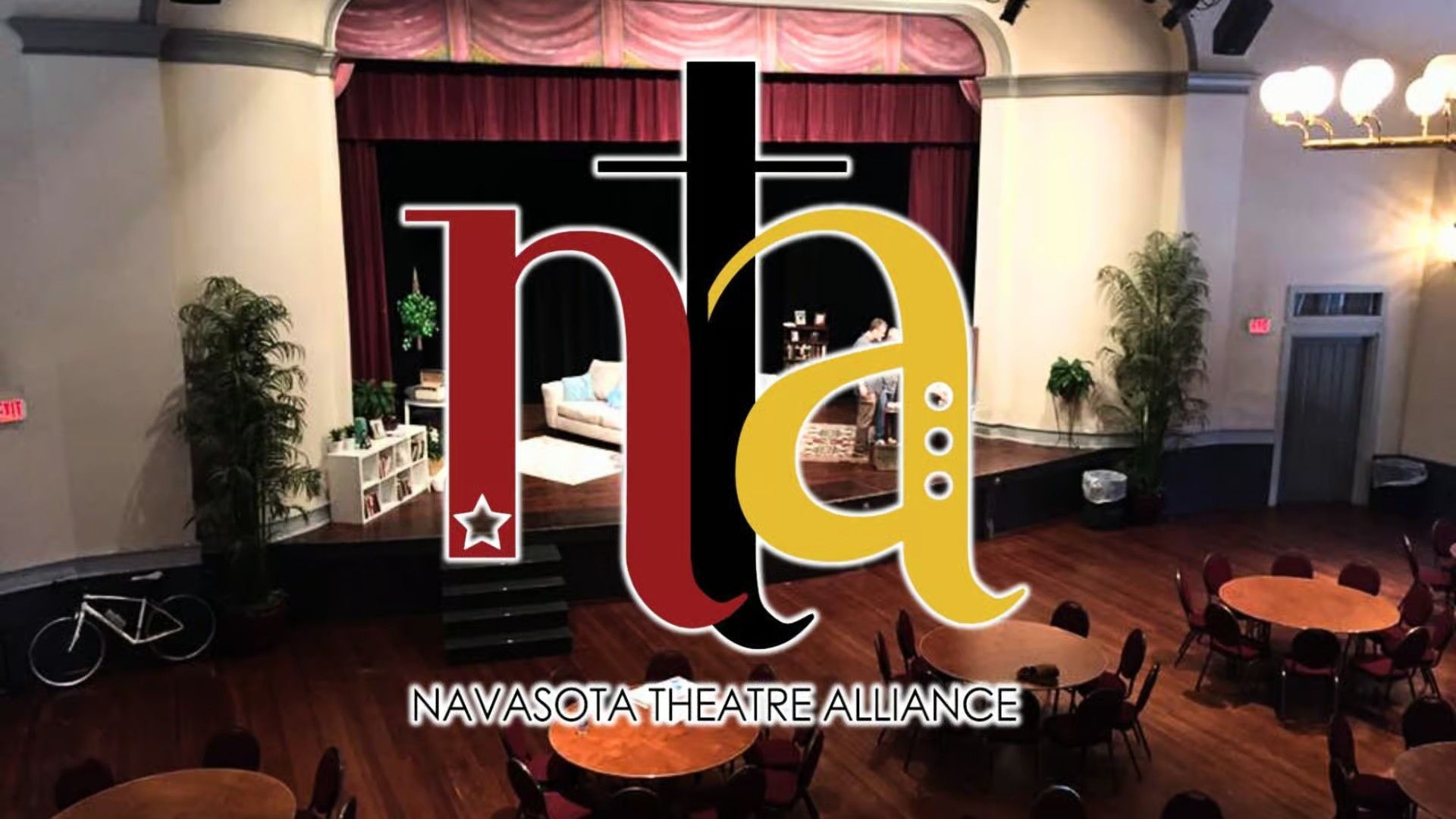Logo for Namsota Theatre Alliance over an indoor stage setup with round dining tables and a wooden floor.