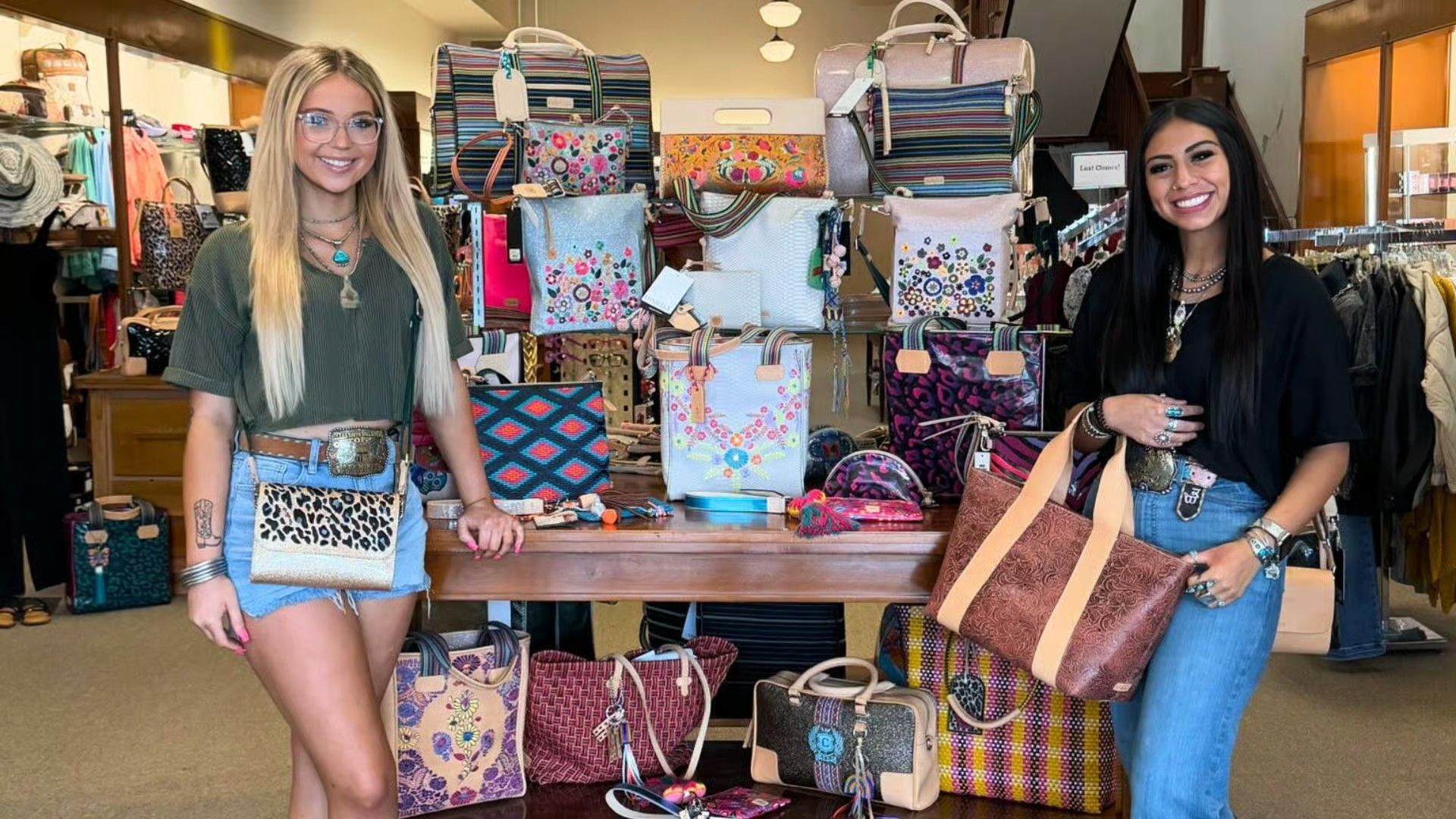 Two people smiling behind a wooden display table filled with assorted embroidered, patterned, and leather handbags.