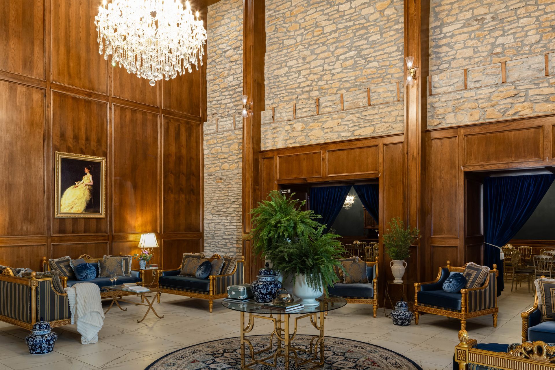 A grand, high-ceilinged lobby with paneled wood walls, stone accents, marble floors, two chandeliers, and plush furniture.