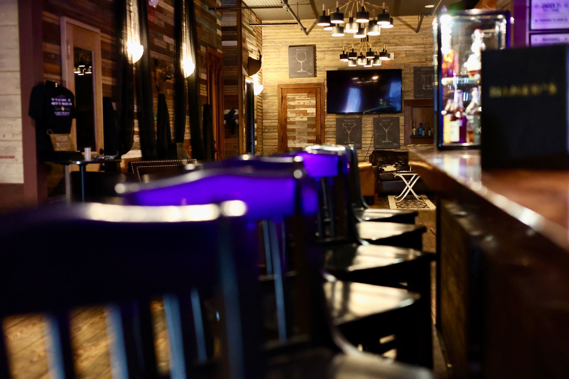 A dimly lit bar interior with a wooden counter, dark chairs, and ambient purple lighting under a warm-toned chandelier.