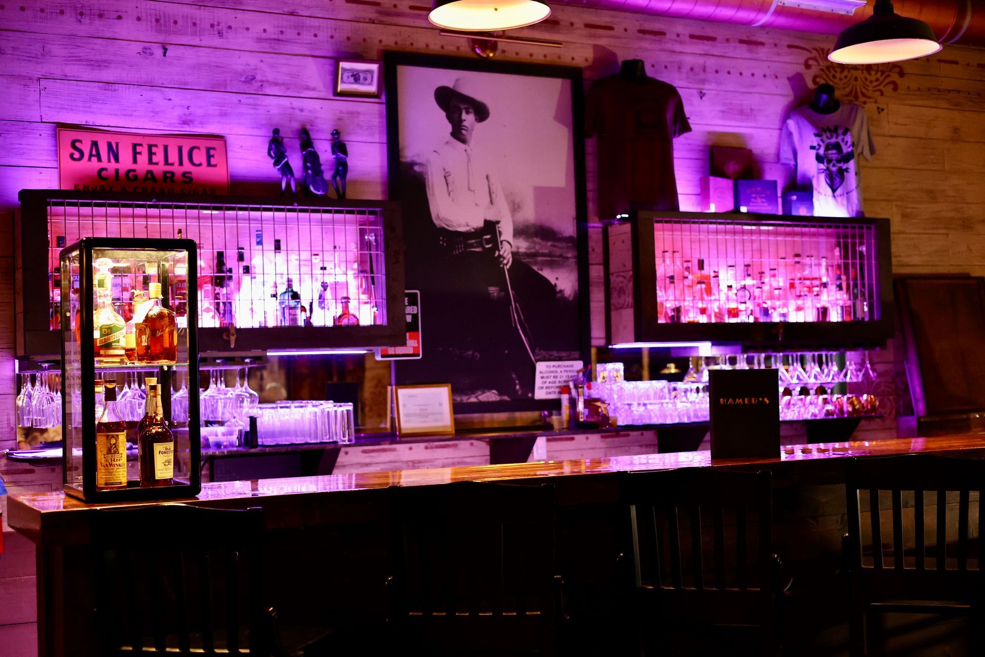 A dimly lit bar with purple lighting, featuring a large framed photo of a person, bottles on display, and wooden counters.