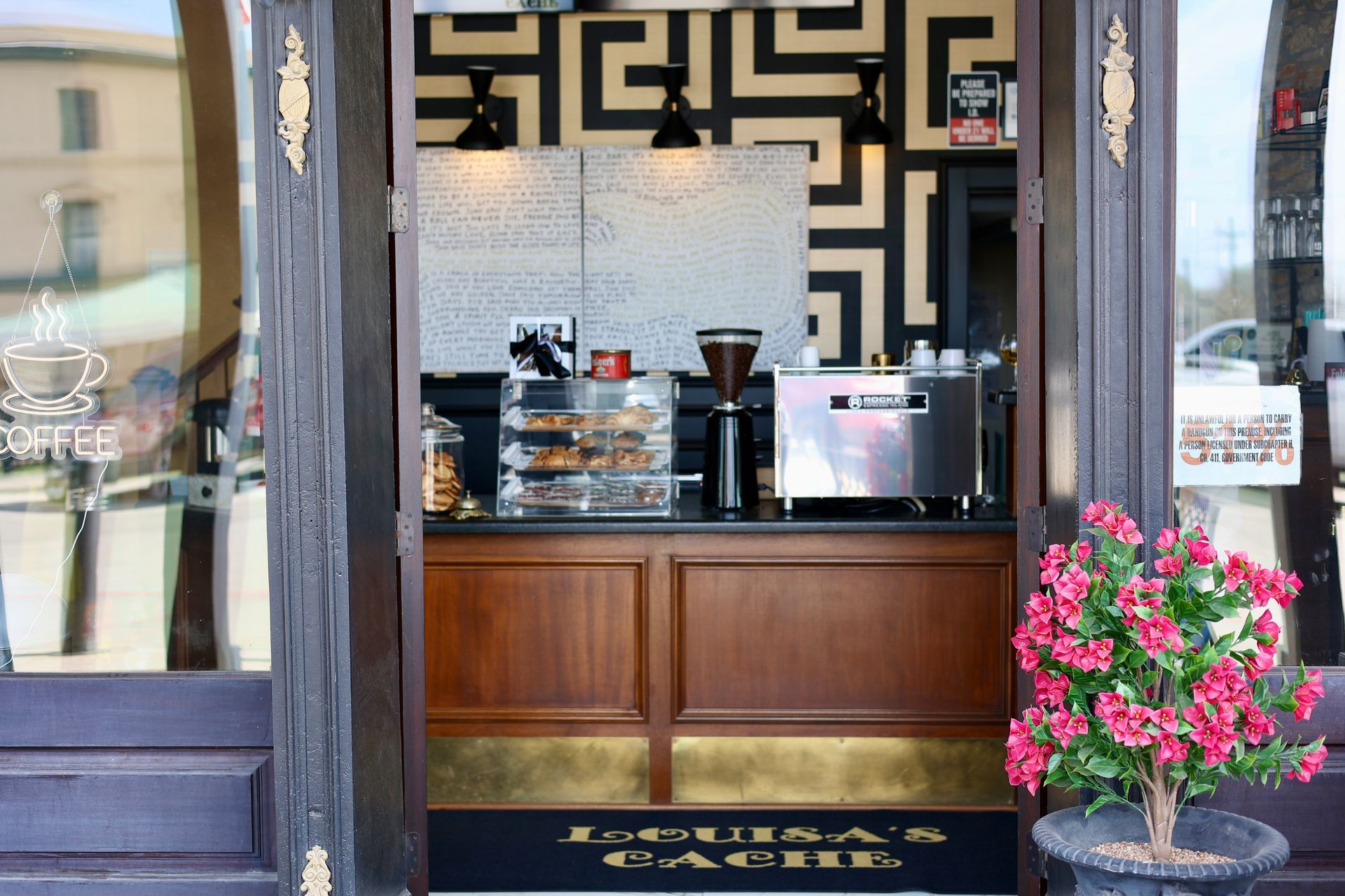 The entrance of Louisa’s Cache, a coffee shop with a wooden counter, a pastry display, espresso machine, and pink flowers.