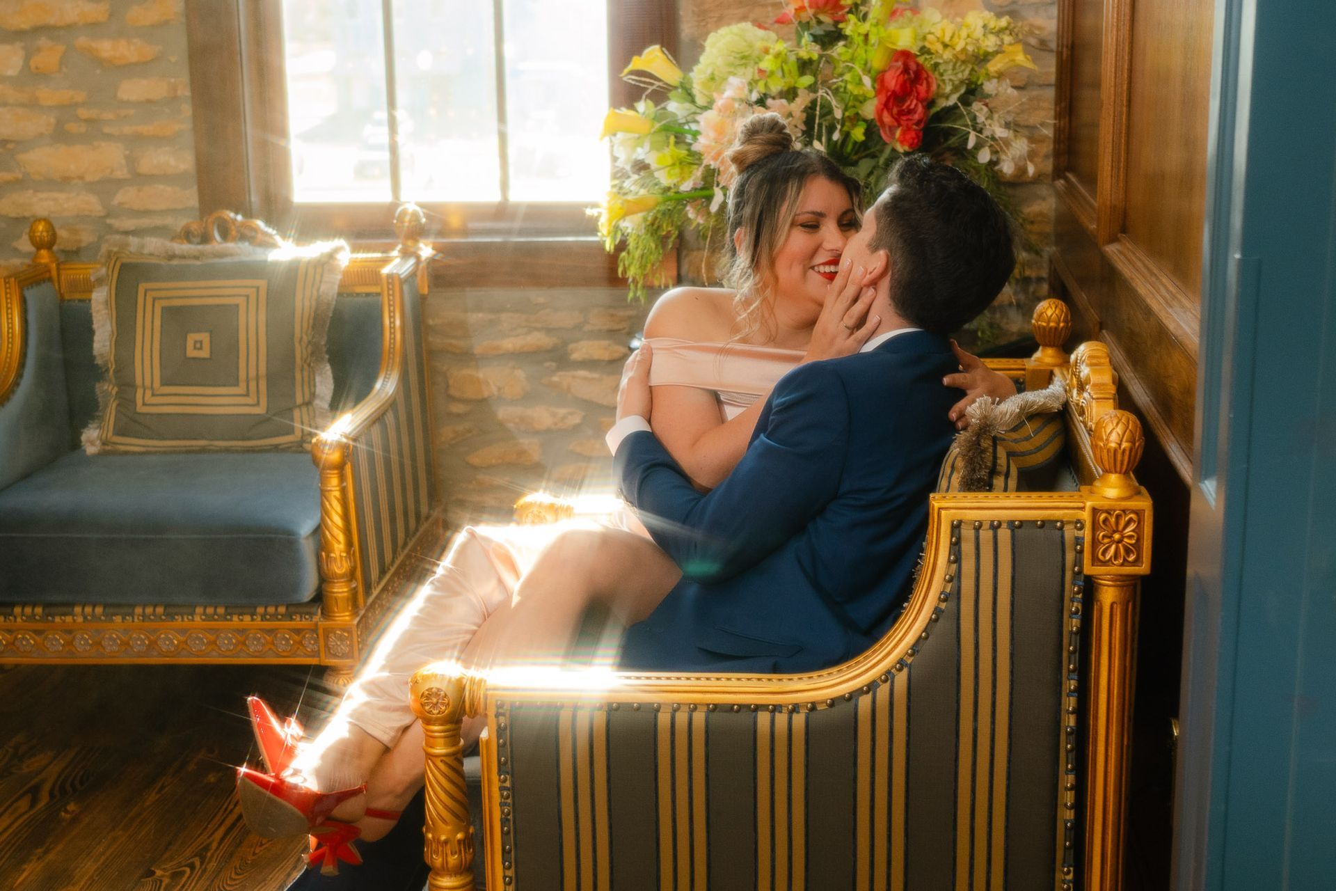 A couple in formal attire embraces while sitting in an ornate, upholstered chair in a bright, rustic room with a window.