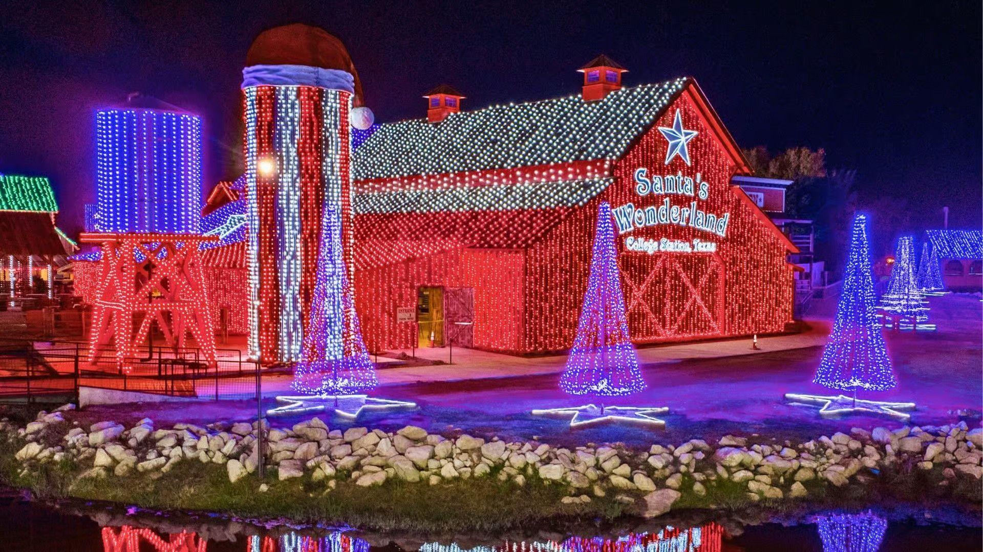 A red barn illuminated with festive Christmas lights next to a blue-lit silo, surrounded by glowing cone-shaped trees.