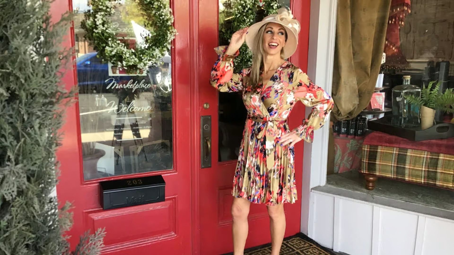 A woman in a floral dress and wide-brim hat smiles while standing outside a storefront with red doors.