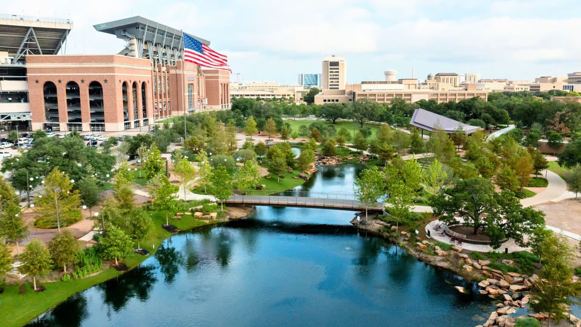 A scenic park with a small pond and wooden footbridge, featuring a large brick stadium with an American flag in the back.