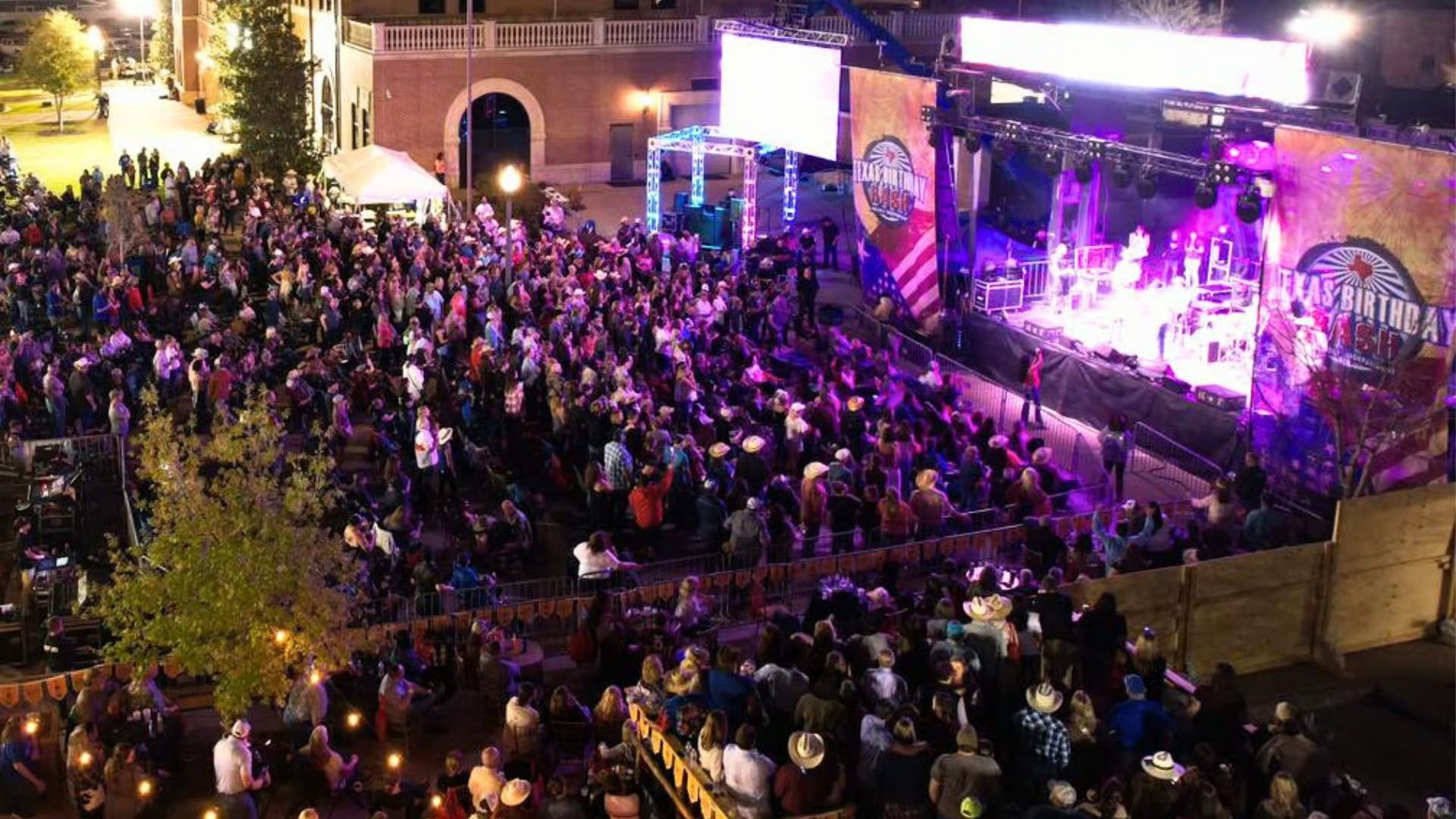 A large crowd gathers in an outdoor plaza at night to watch a live concert on a brightly lit stage.