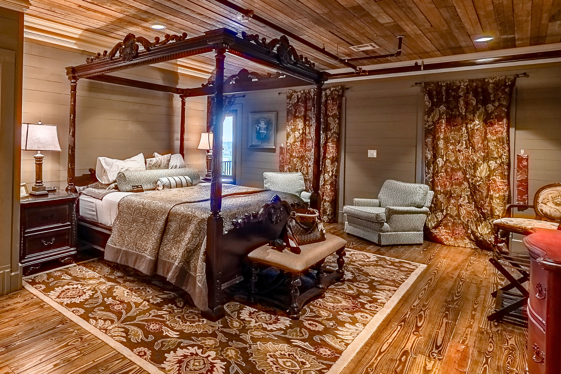 A rustic bedroom featuring a dark wood four-poster bed, ornate patterned rug, wood-paneled ceiling, and floral curtains.