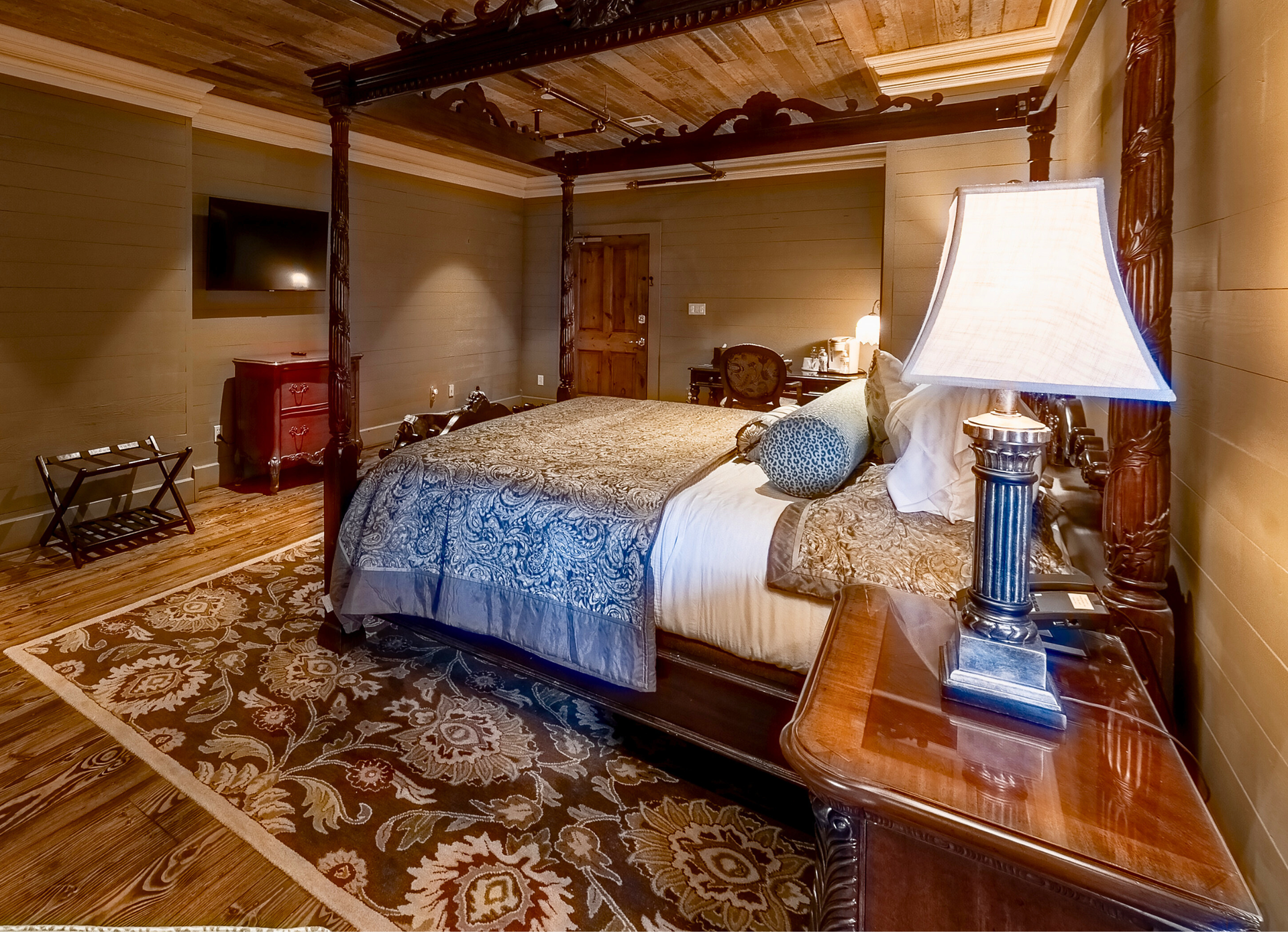 A master bedroom with a large dark wood four-poster bed, patterned rug, lamp, and television on a dark olive wall.