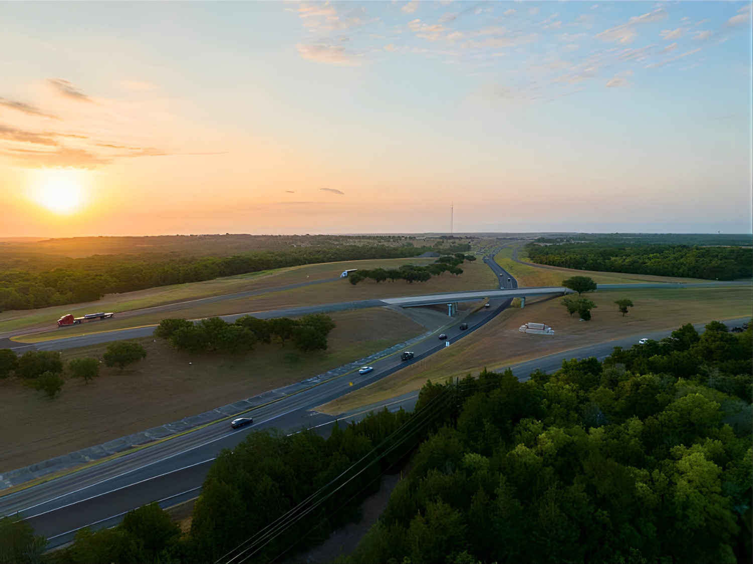 An aerial view of a multi-lane highway winding through a rural, grassy landscape during a golden sunrise.