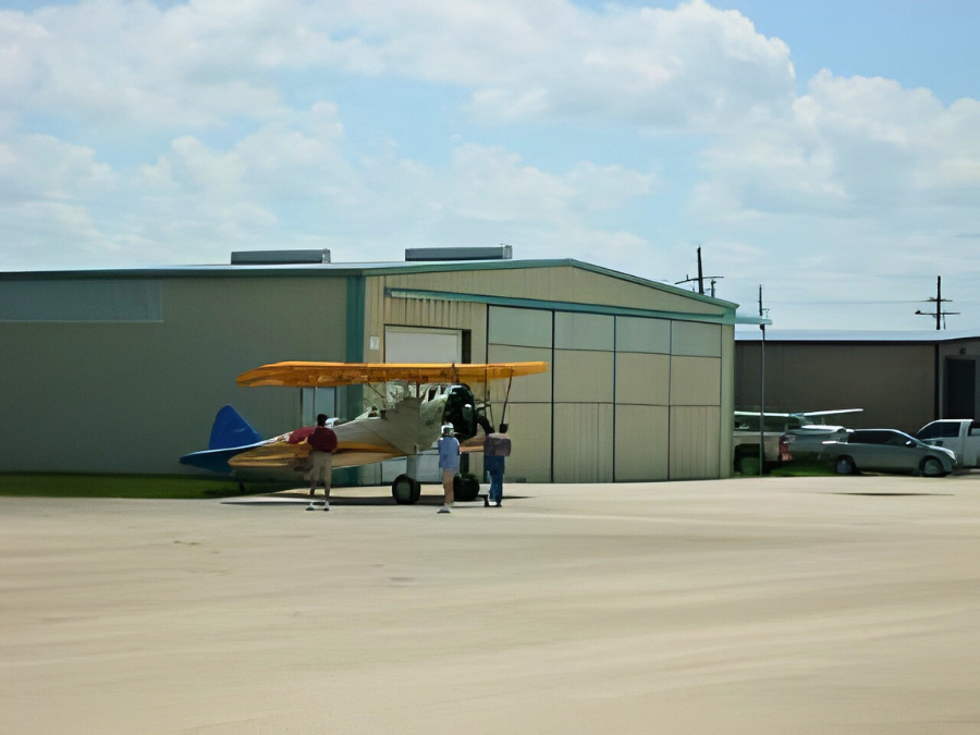 A yellow biplane parked outside a hangar at an airfield with three people standing near it.
