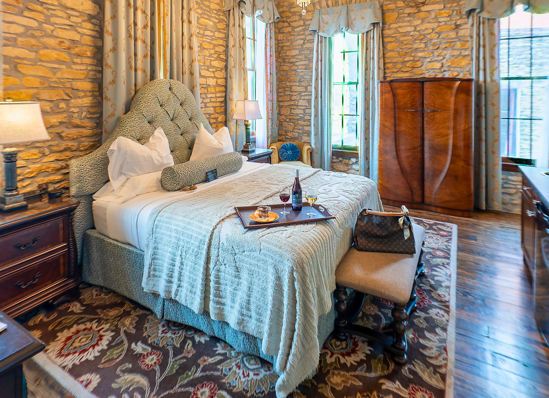 A rustic bedroom with stone walls, a patterned bed with a breakfast tray, wood furniture, and a floral area rug.