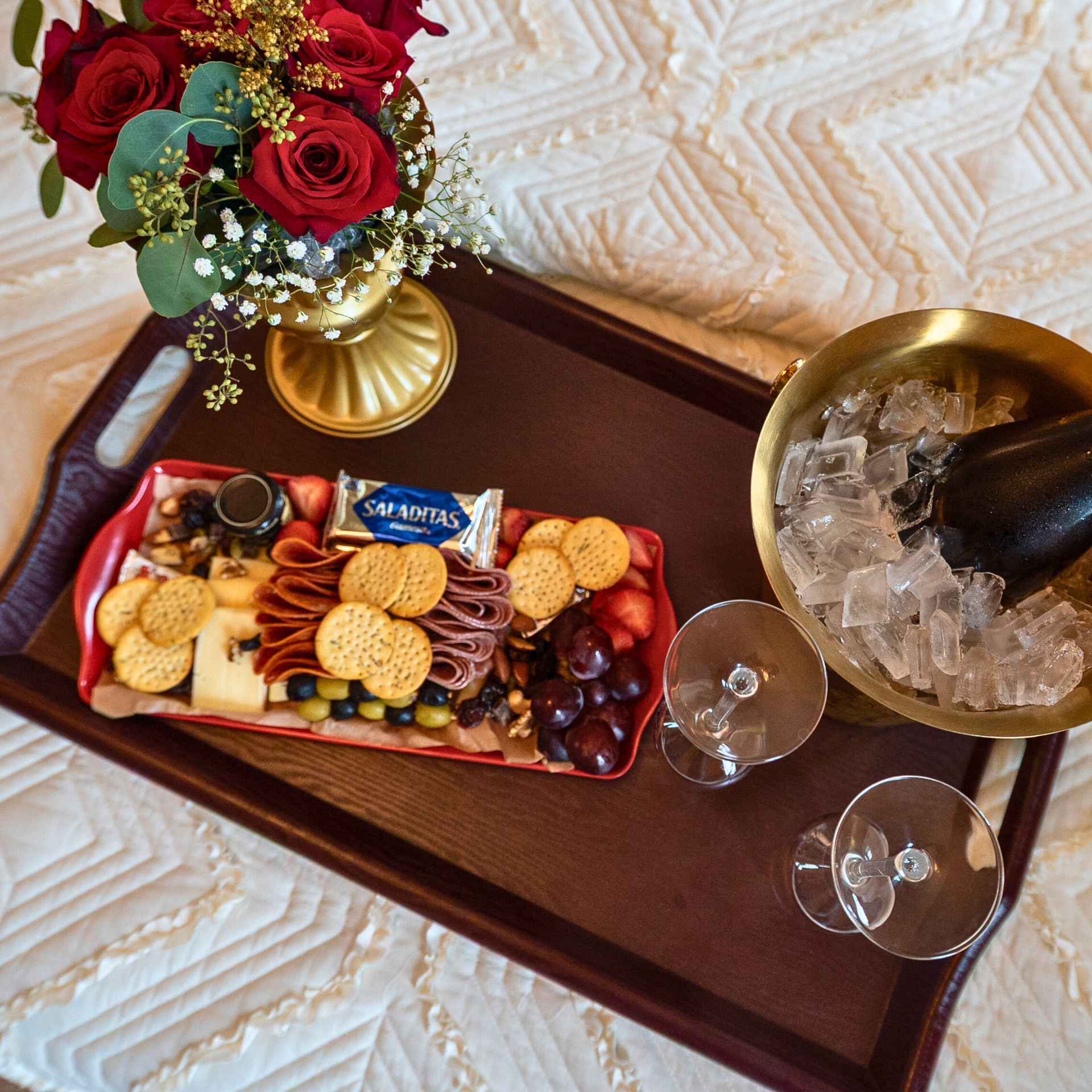 A dark wood tray on a quilted surface holds a charcuterie board, a vase of red roses, two glasses, and an ice bucket.