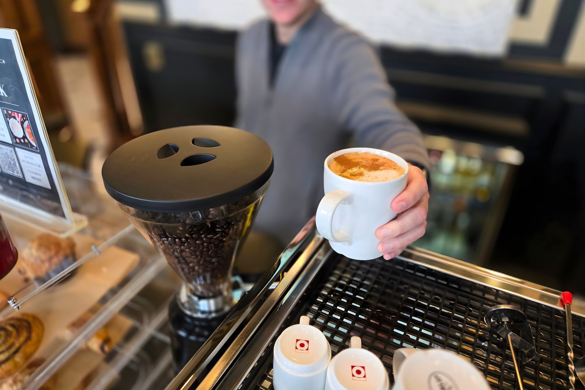A person behind a counter hands a steaming white mug of coffee to a customer in a cafe.
